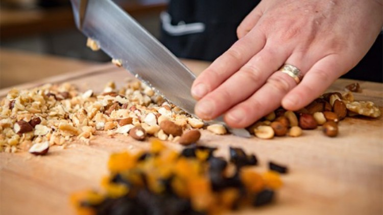 Eine Hand hält ein Messer und hackt Nüsse auf einem Holzbrett, im Vordergrund liegen getrocknete Früchte.