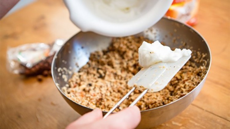 Eine Schüssel mit einer körnigen Mischung, daneben wird mit einem Teigschaber weiße Creme aus einer Schüssel hinzugefügt.