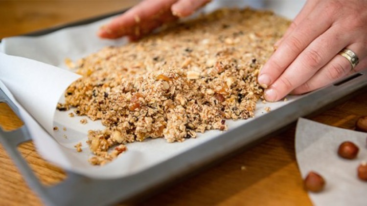 Hände formen auf einem mit Backpapier ausgelegten Backblech eine Masse aus Haferflocken und Nüssen, daneben liegen Haselnüsse.