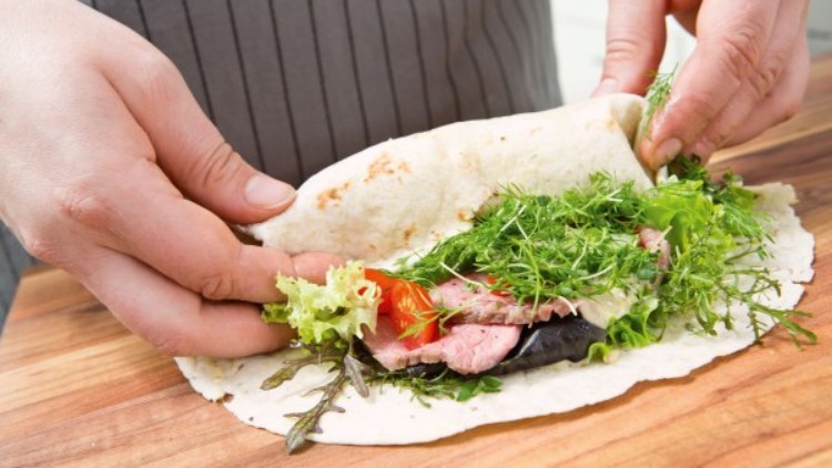 Hände rollen einen Tortilla-Wrap mit Salat, frischen Kräutern, Tomatenscheibe und Fleisch auf einem Holzuntergrund ein.
