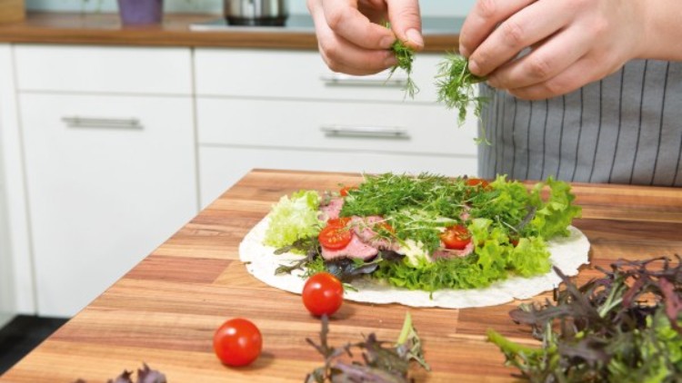 Person in Schürze belegt ein Wrap mit frischem Salat, Kräutern, Tomaten und weiteren Zutaten in einer hellen Küche.
