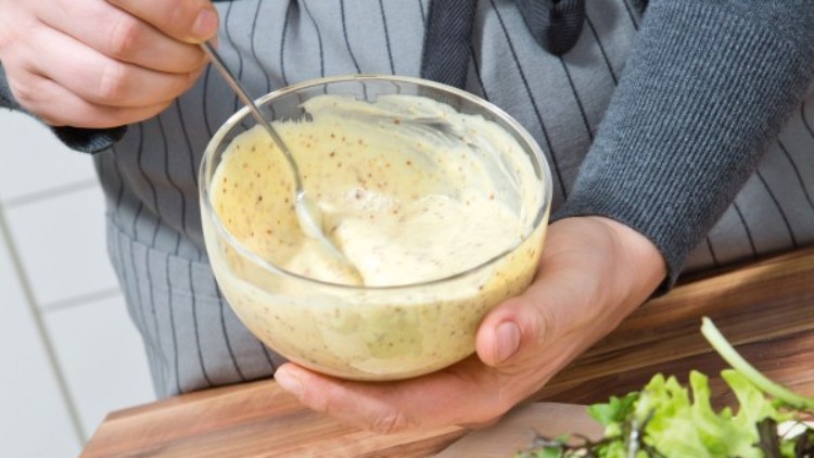 Person in grauer Schürze hält eine Schüssel mit Senf-Dressing, daneben liegt Salat mit Gurken und Tomaten auf einem Schneidebrett.