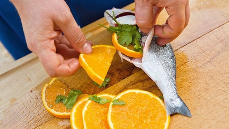 Eine Person füllt einen offenen Fisch mit frischen Orangenscheiben und grünem Koriander auf einem Holzbrett.