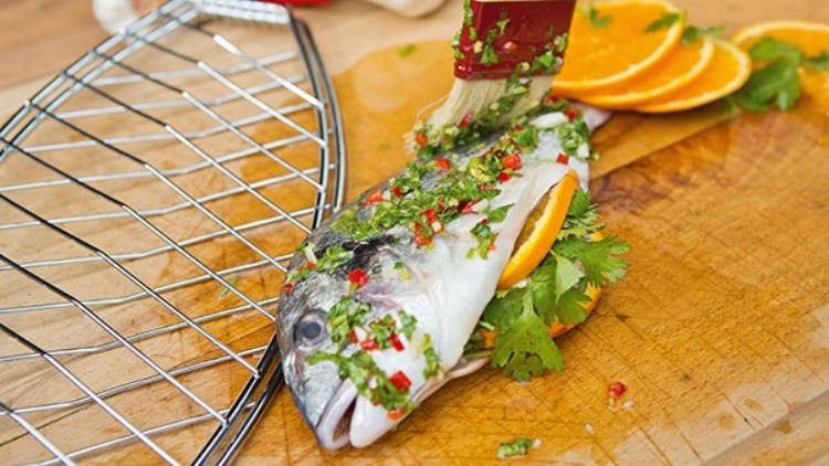 Frischer Fisch mit Kräutermarinade, Orangen und Koriander wird auf einem Holzbrett zubereitet, daneben ein Grillgitter.