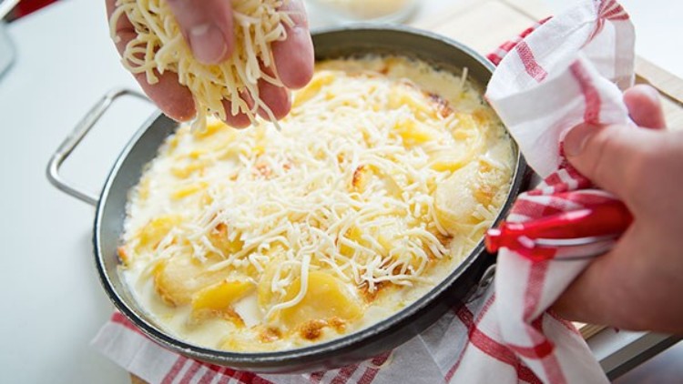 Eine runde Pfanne mit überbackenen Kartoffelscheiben, darüber werden geriebener Käse gestreut und ein rotes Küchentuch gehalten.