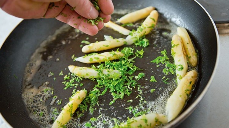 Weiße Spargelstangen werden in einer Pfanne mit Butter und gehackten Kräutern zubereitet, eine Hand streut Kräuter darüber.