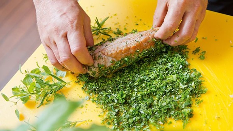 Hände rollen ein Stück Fleisch in gehackten frischen Kräutern auf einer gelben Unterlage.