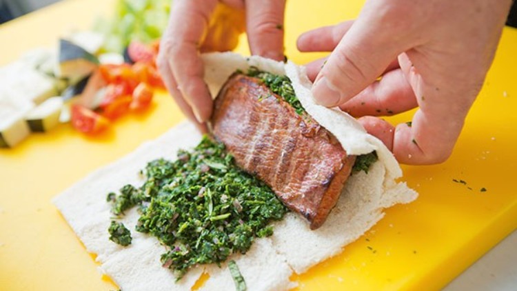Hände rollen ein Sandwich mit gegrilltem Fleisch und gehackten Kräutern auf einer gelben Unterlage, Gemüse im Hintergrund.