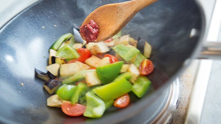 Gemüse wie Paprika, Aubergine und Tomaten wird in einer schwarzen Pfanne auf einem Herd mit einem Holzlöffel zubereitet.