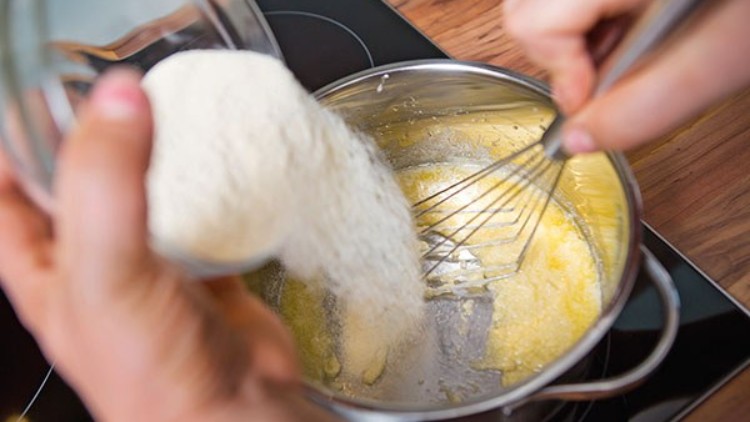 Mehl wird aus einem Glas in einen Topf mit geschmolzener Butter geschüttet, während eine Hand mit einem Schneebesen rührt.