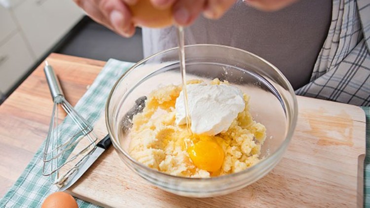 Hand schlägt ein Ei in eine Glasschüssel mit Teig und weißer Creme, daneben ein Schneebesen und ein Geschirrtuch.