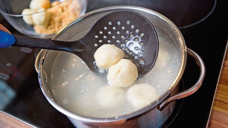 Ein schwarzer Schaumlöffel hebt einen Knödel aus einem Topf mit kochendem Wasser, im Hintergrund steht eine Schüssel mit weiteren Knödeln.