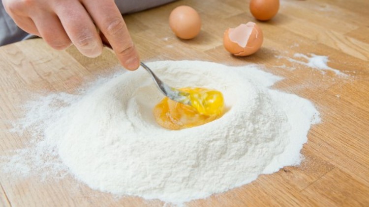 Eine Person vermischt mit einer Gabel Eigelb in einem Mehlhügel auf einer Holzarbeitsfläche, daneben liegen zwei Eier und eine Eierschale.