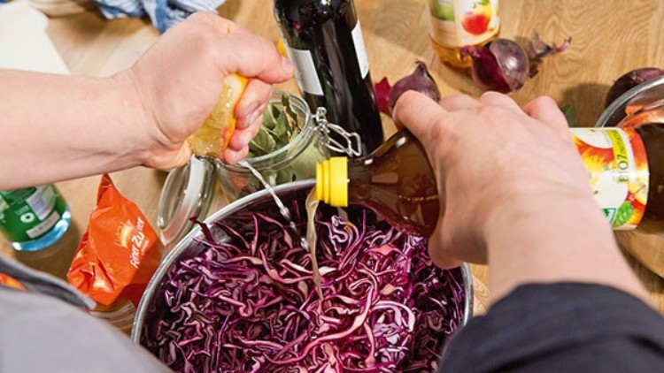Geriebenes Rotkraut in einem Topf, in das Zitronensaft gedrückt und Essig aus einer gelben Flasche hinzugegeben wird.