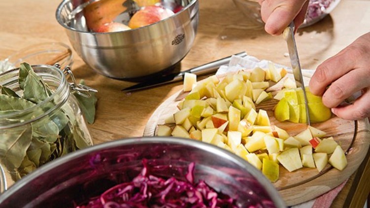 Eine Hand schneidet auf einem Holzbrett gelbe Äpfel klein, daneben Rotkraut, Lorbeerblätter und eine Schüssel mit Äpfeln.