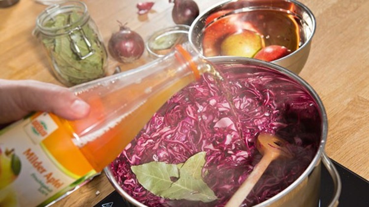 Eine Hand gießt milden Apfelessig aus einer Flasche in einen Topf mit Rotkraut, Lorbeerblatt und Kochlöffel auf einem Herd.