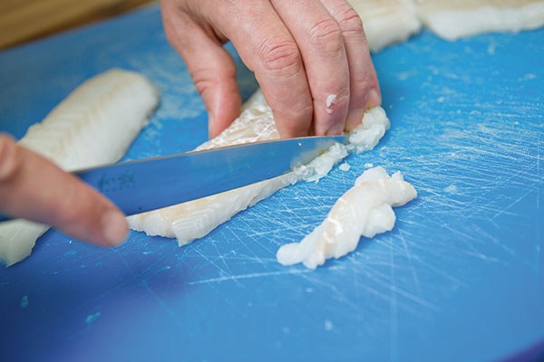 Hände schneiden weißen Fisch mit einem Messer auf einem blauen Schneidebrett in kleine Stücke.