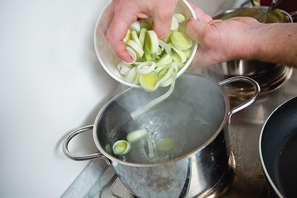 In einen Kochtopf mit kochendem Wasser wird geschnittenes Gemüse aus einer Schale geschüttet, darunter Lauchringe.