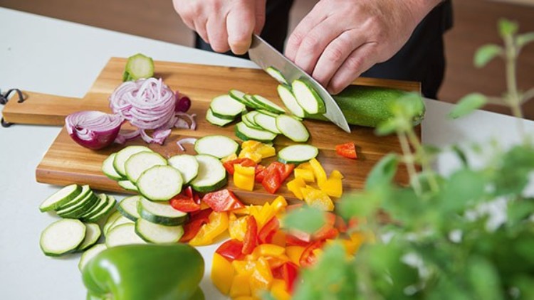 Hände schneiden mit einem Messer Zucchini auf einem Holzbrett, darauf auch Paprika, Zwiebeln und weitere Zucchinischeiben.