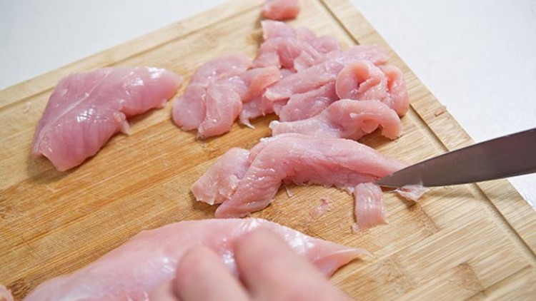 Eine Hand schneidet rohes Hühnerfleisch mit einem Messer auf einem hellen Holzbrett in schmale Streifen.