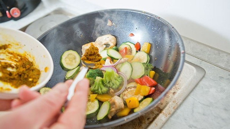 Eine Person gibt mit einem Löffel gelbe Currypaste in eine Wokpfanne mit Gemüse wie Zucchini, Brokkoli und Paprika.