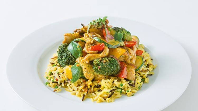 Ein weißer Teller mit Gemüse-Curry und Brokkoli auf gelbem Reis, garniert mit frischen Kräutern.