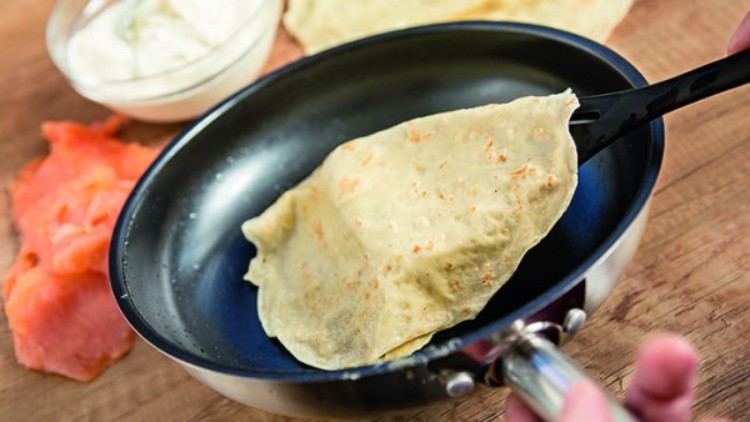Ein dünner, hellbraun gebackener Pfannkuchen wird in einer schwarzen Pfanne mit einem Pfannenwender gewendet.