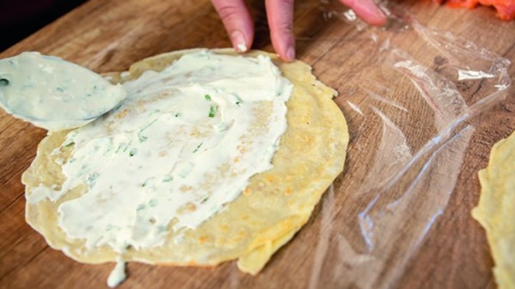 Eine Hand streicht Frischkäse mit Kräutern auf einen dünnen Pfannkuchen auf einem Holzbrett, daneben liegt geräucherter Lachs.