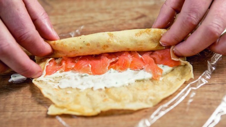 Eine Person rollt einen Crêpe mit Kräutercreme und Räucherlachs auf einer durchsichtigen Folie zusammen.