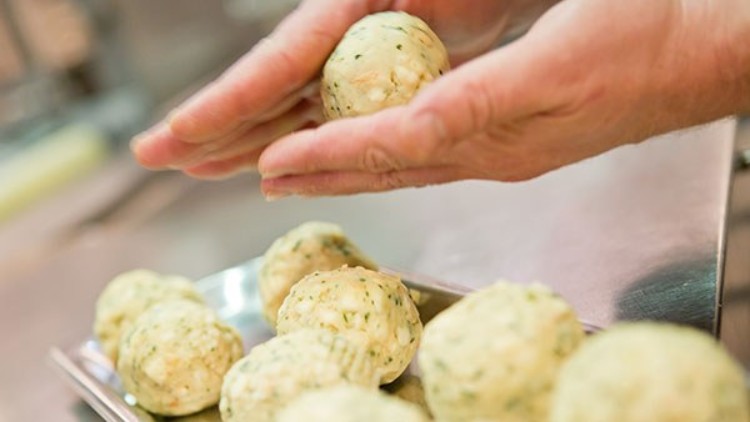 Zwei Hände formen runde, roh aussehende Knödel mit Kräutern, weitere Knödel liegen auf einem Tablett im Hintergrund.