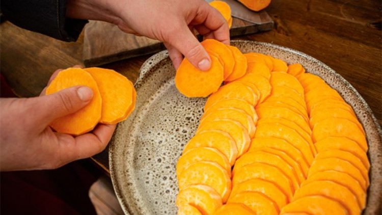 Zwei Hände legen orangefarbene Süßkartoffelscheiben auf einen grauen Teller, der auf einem Holztisch steht.