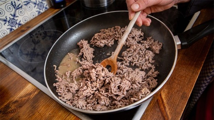 Eine Hand rührt mit einem Holzlöffel in einer Bratpfanne, in der angebratenes Hackfleisch auf einem Herd liegt.
