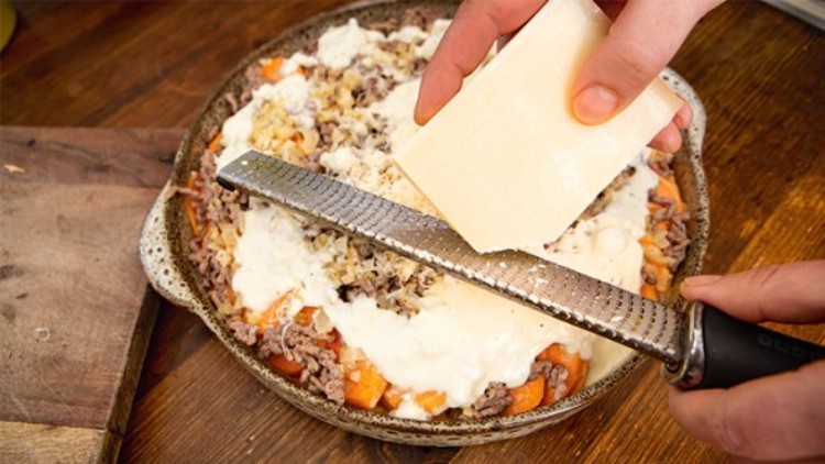 Eine Hand reibt hellen Käse mit einer feinen Reibe über eine mit Hackfleisch, Gemüse und Soße gefüllte Auflaufform.