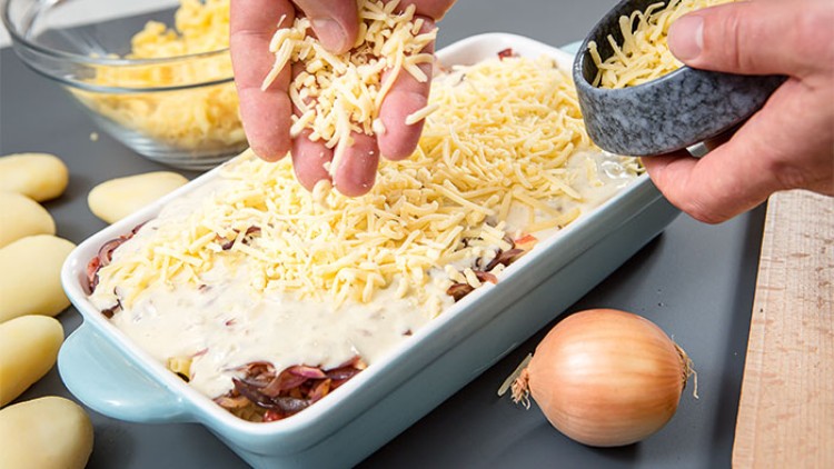 Eine rechteckige Auflaufform wird mit geriebenem Käse bestreut, daneben liegen Kartoffeln, eine Zwiebel und eine Schüssel Käse.