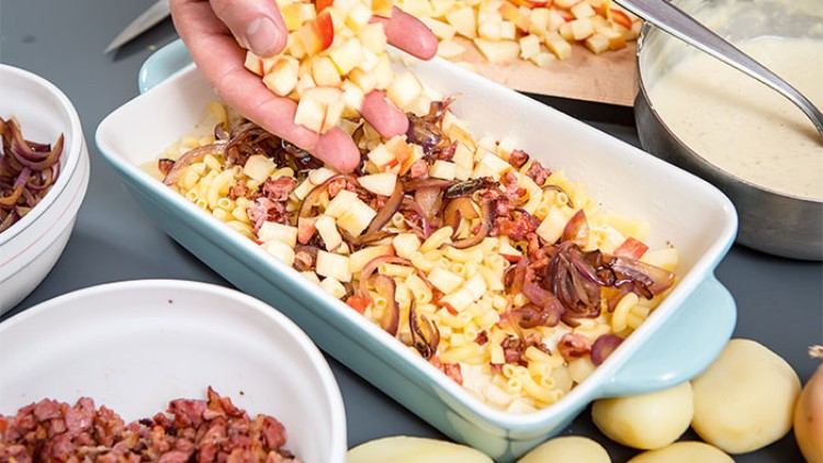 Eine Hand streut gewürfelte Apfelstücke über eine Auflaufform mit Nudeln, Speck und Zwiebeln, daneben liegen Kartoffeln und Zutaten.