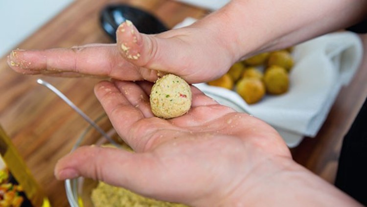 Eine Person formt mit den Händen eine kleine Kugel aus Falafel-Teig, im Hintergrund eine Glasschüssel und fertige Falafel.