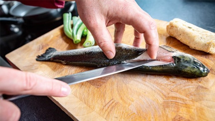 Eine Hand filetiert mit einem Messer einen rohen Fisch auf einem Holzbrett, daneben Bohnen und eine Teigrolle.
