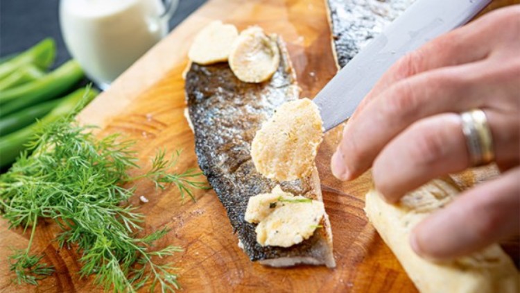 Eine Hand streicht mit einem Messer Kräuterbutter auf ein Stück Fischfilet mit Haut, daneben liegen frischer Dill und Frühlingszwiebeln.
