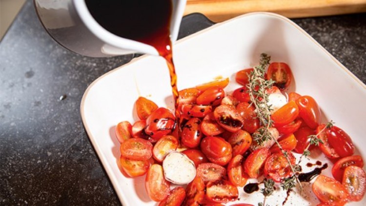Kleine Tomaten, Mozzarella und frischer Thymian in einer weißen Form werden mit dunklem Balsamico aus einem Krug übergossen.