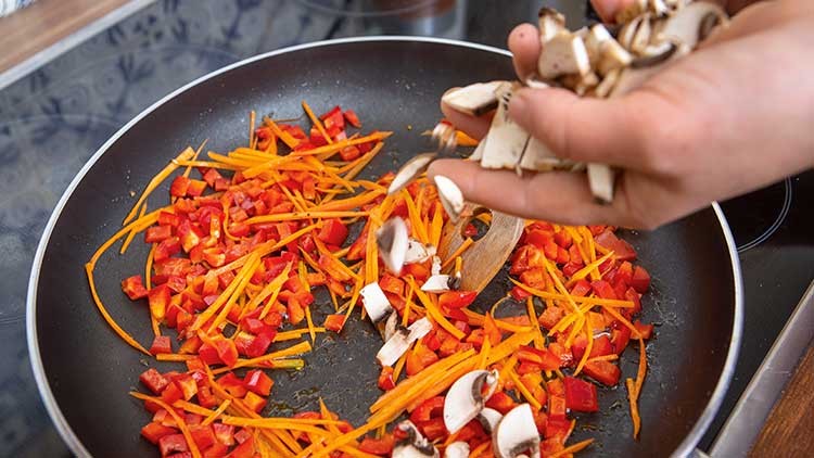 In einer schwarzen Pfanne werden Karottenstreifen, Paprikastücke und Champignons von einer Hand hinzugefügt und angebraten.