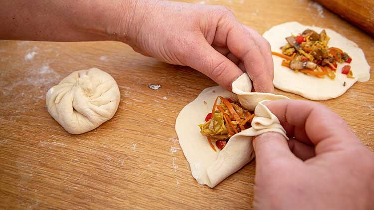 Zwei Hände falten Teig mit Gemüsefüllung, ein fertig geschlossener Teigling liegt daneben auf einer Holzoberfläche.