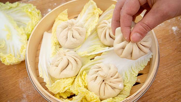 Gedämpfte Teigtaschen liegen auf hellgrünen Chinakohlblättern in einem Bambusdämpfer, eine Hand legt eine weitere Teigtasche hinein.