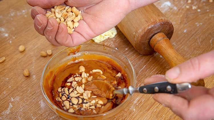 Eine Hand streut gehackte Erdnüsse in eine Schüssel mit Karamellcreme, daneben liegt ein Nudelholz auf einer bemehlten Holzfläche.