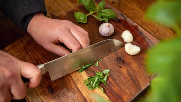 Hände schneiden frische grüne Kräuter mit einem Messer auf einem Holzbrett, daneben Knoblauch und Basilikumblätter.