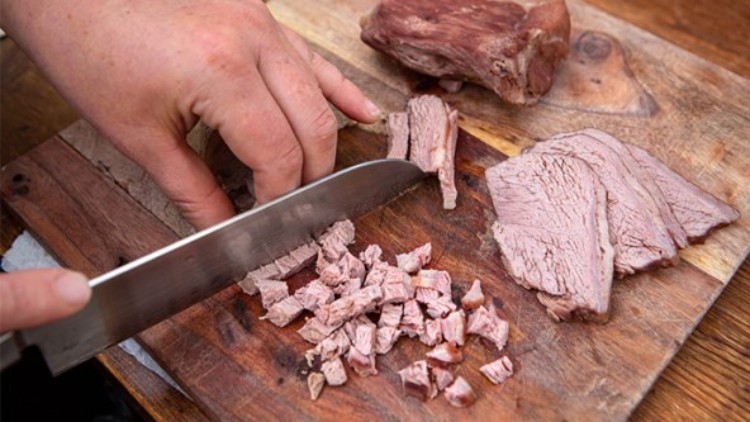 Eine Hand schneidet mit einem Messer gekochtes Rindfleisch auf einem Holzbrett in kleine Würfel, daneben liegen Fleischstücke.