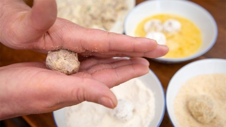 Zwei Hände formen eine ungebratene Frikadelle, daneben Schüsseln mit Mehl, verquirltem Ei und Paniermehl.