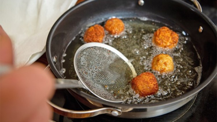 Vier goldbraune, runde Bällchen werden in heißem Öl in einer beschichteten Pfanne frittiert, eine Schaumkelle ist im Einsatz.