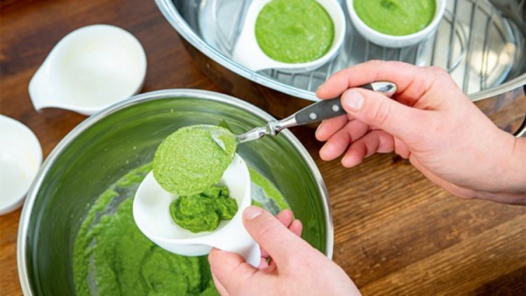 Eine Person füllt grüne Masse aus einer Schüssel mit einem Löffel in ein weißes Förmchen, weitere Förmchen sind im Dampfgarer.