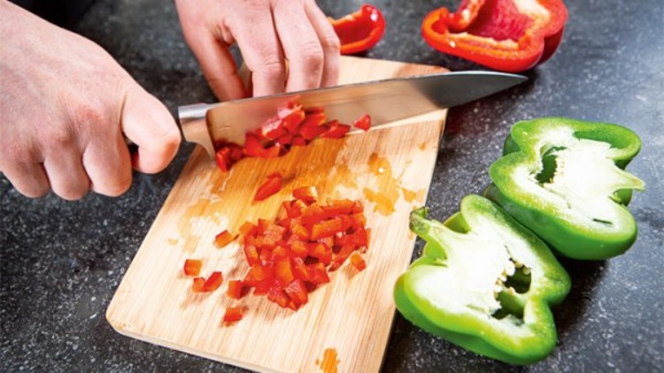 Hände schneiden rote Paprika auf einem Holzbrett in Würfel, daneben liegen halbe grüne und rote Paprika auf schwarzer Arbeitsplatte.