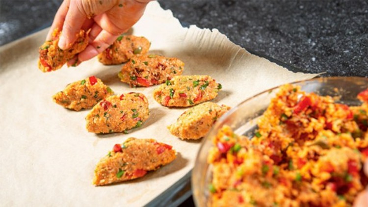 Eine Hand formt mit einem Löffel vegetarische Teigportionen mit Gemüse und legt sie auf ein mit Backpapier belegtes Blech.